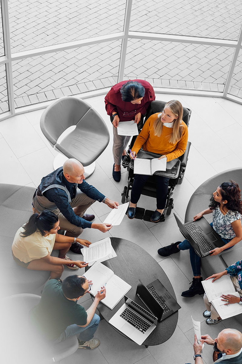 A top-down view of people of various diversities and backgrounds sitting around a low table in the middle of a meeting.
